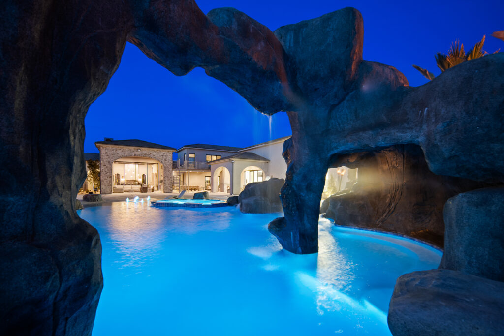 Luxury custom home pool area with rock formations and illuminated water features, showcasing a tranquil outdoor living space in Ivins, Utah.