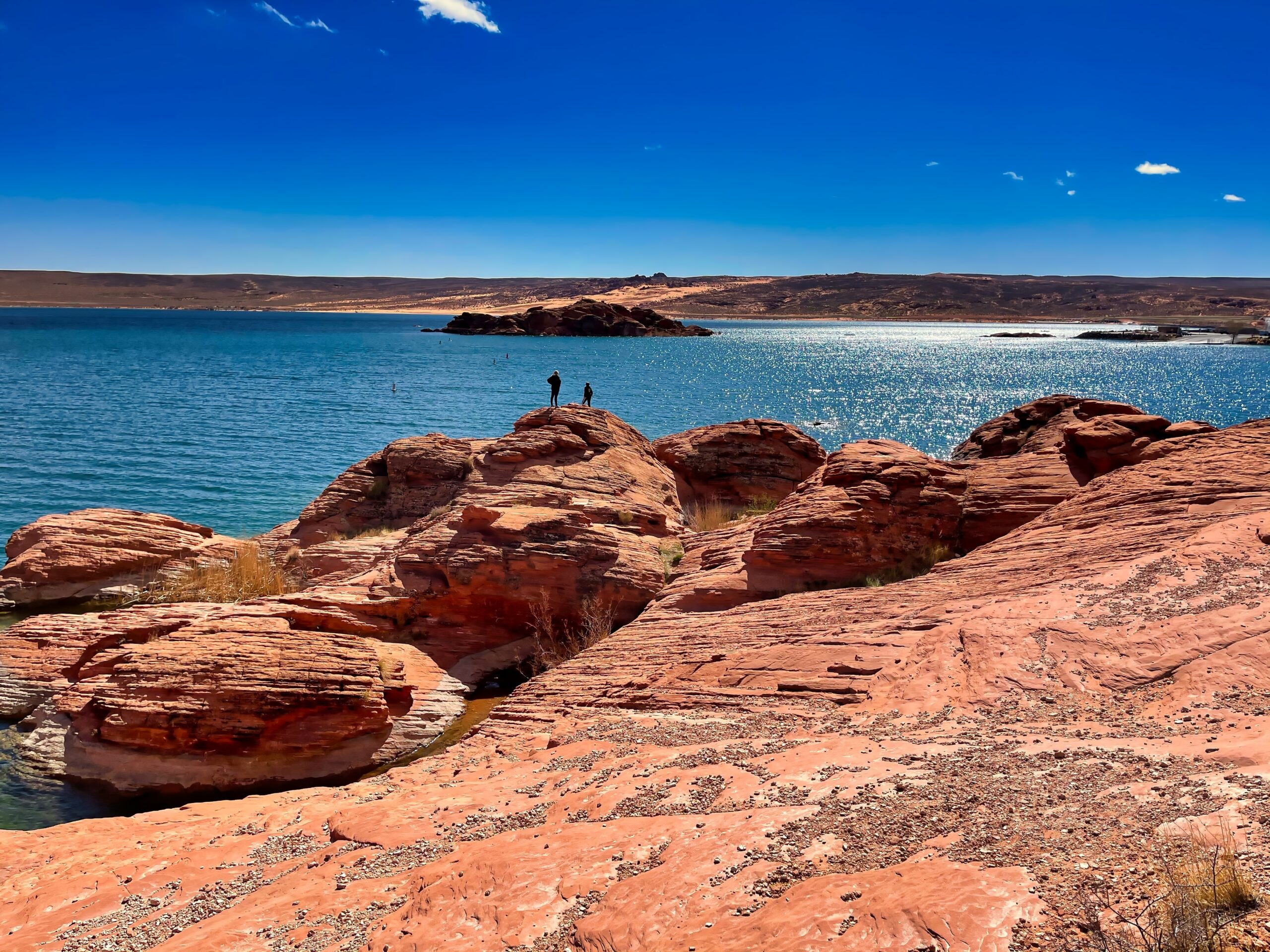 Scenic view of red rock formations and a tranquil lake in Hurricane, Utah, with two figures standing on the rocks, highlighting the area's natural beauty and outdoor recreational opportunities.