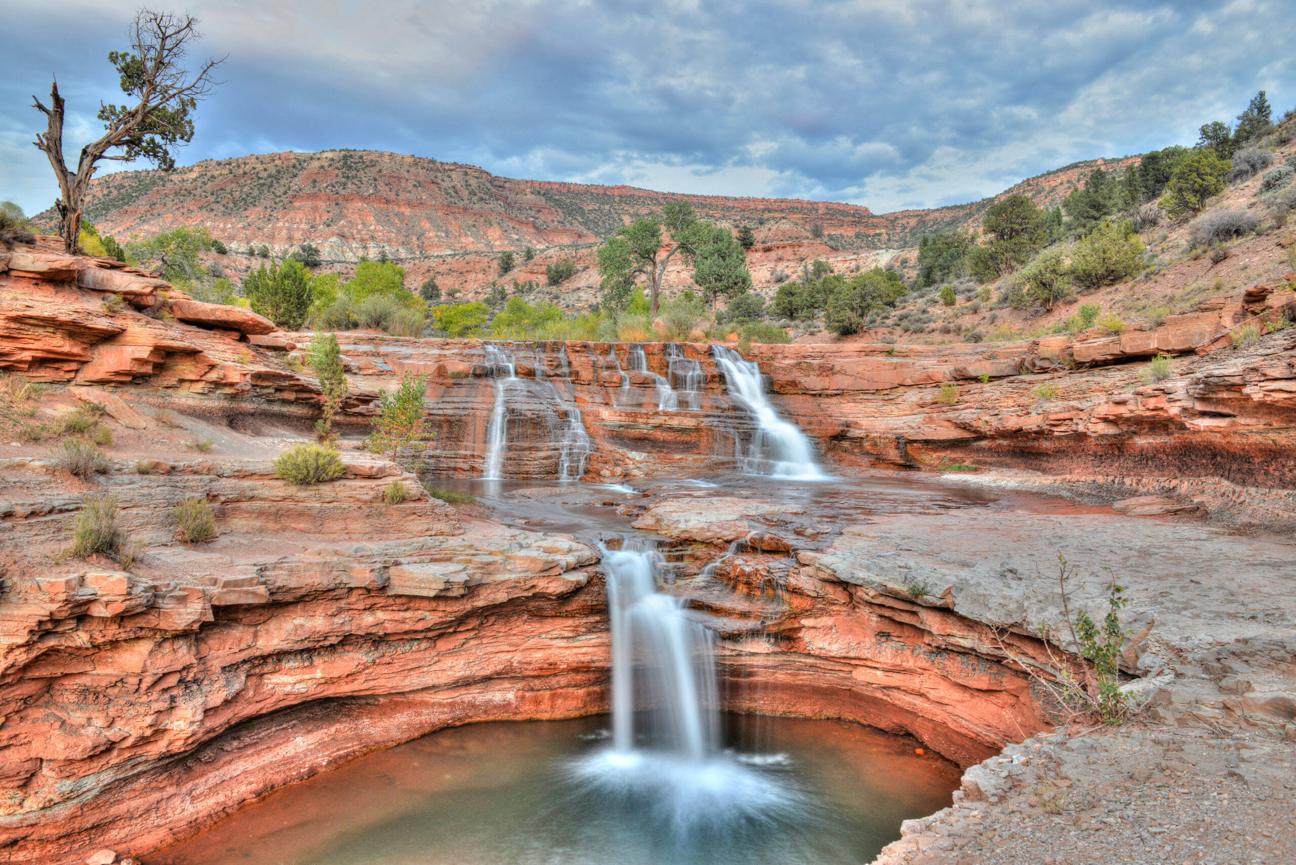 Scenic waterfall cascading over red rock formations in Toquerville, Utah, surrounded by lush greenery and rugged hills, emphasizing the area's natural beauty and outdoor recreational appeal.
